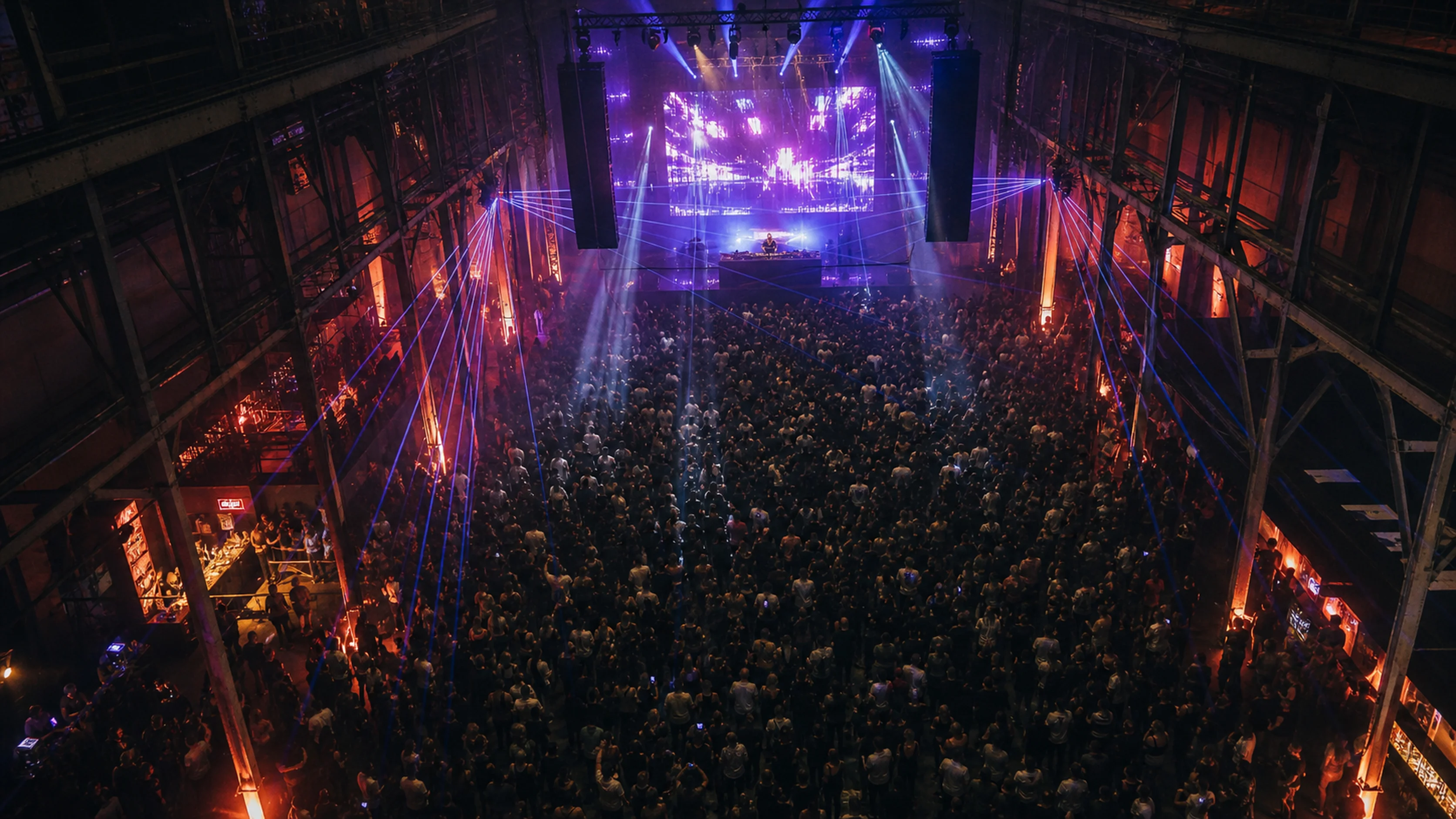 Aerial view of a massive indoor warehouse event — LED wall main stage, lasers crossing the room, packed crowd